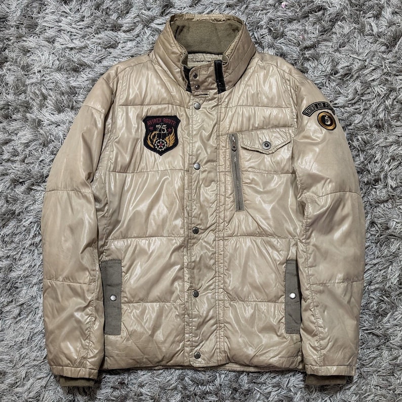 Peut inclure: Veste matelass&eacute;e beige avec un col montant, fermeture &eacute;clair et boutons. La veste comporte plusieurs poches et &eacute;cussons avec du texte et des embl&egrave;mes. La veste est de couleur neutre et semble &ecirc;tre faite d'un mat&eacute;riau brillant.