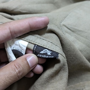 May include: A tan-coloured garment featuring a black fabric tag with the white text AVIREX and a stylised A logo. A white care label is visible nearby, displaying the text NO. 611579, alongside a dark button attached to the textured fabric.