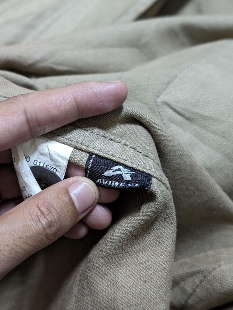 May include: A tan-coloured garment featuring a black fabric tag with the white text AVIREX and a stylised A logo. A white care label is visible nearby, displaying the text NO. 611579, alongside a dark button attached to the textured fabric.