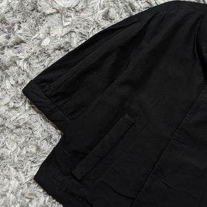 May include: A black shirt with a textured, crinkled fabric finish, featuring short sleeves with cuffed hems and a vertical pocket detail on the front. The garment has a structured collar and a clean, solid black color throughout the design.