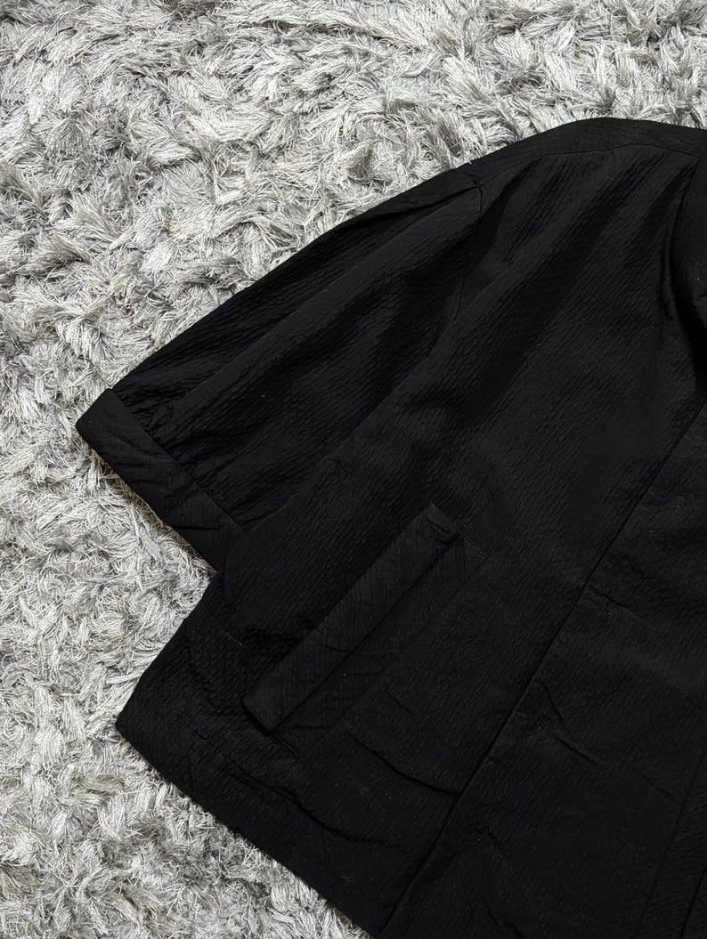 May include: A black shirt with a textured, crinkled fabric finish, featuring short sleeves with cuffed hems and a vertical pocket detail on the front. The garment has a structured collar and a clean, solid black color throughout the design.
