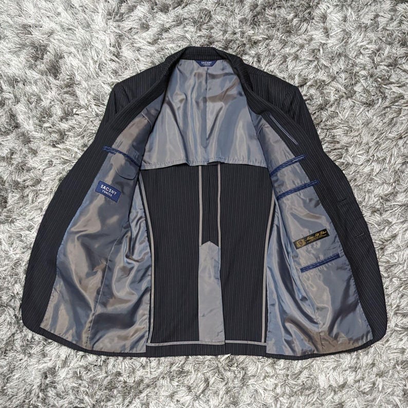 May include: A dark gray pinstripe suit jacket with the interior visible. The jacket has a gray lining, multiple pockets, and a label that reads "SACSNY". The jacket is open, displaying the interior construction and design.