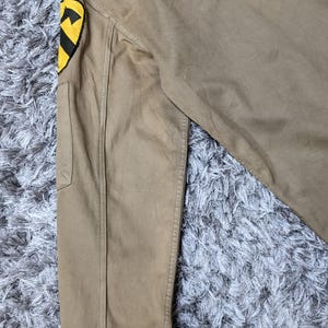 May include: A tan long-sleeve jacket featuring a prominent yellow and black shield-shaped patch on the upper sleeve. The patch displays a black horse head silhouette against a yellow background, with a black horizontal stripe across the centre.