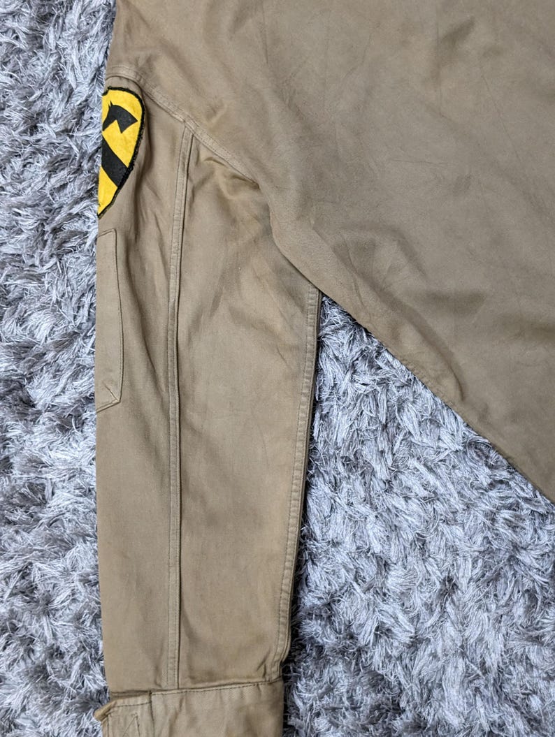 May include: A tan long-sleeve jacket featuring a prominent yellow and black shield-shaped patch on the upper sleeve. The patch displays a black horse head silhouette against a yellow background, with a black horizontal stripe across the centre.