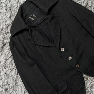 May include: A black blazer with a wide collar and three buttons on the front. The interior label features the text Y's. The garment has long sleeves and two small flap pockets at the waist, constructed with a structured fit and visible stitching.