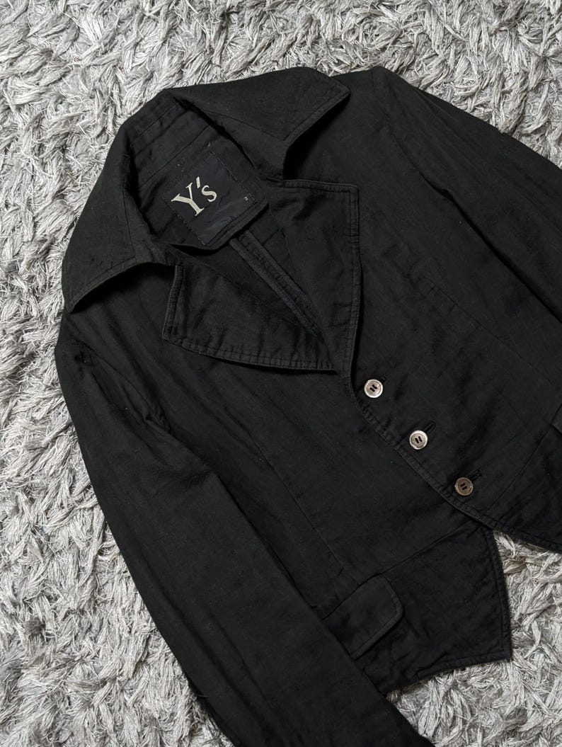 May include: A black blazer with a wide collar and three buttons on the front. The interior label features the text Y's. The garment has long sleeves and two small flap pockets at the waist, constructed with a structured fit and visible stitching.