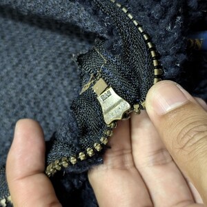 May include: Close-up of a dark blue fleece jacket with a brass zipper. The zipper is partially open, revealing the textured fabric and a small blue label. The jacket appears to be made of a thick, warm material.