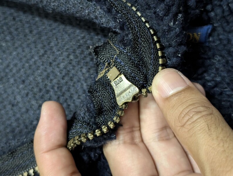 May include: Close-up of a dark blue fleece jacket with a brass zipper. The zipper is partially open, revealing the textured fabric and a small blue label. The jacket appears to be made of a thick, warm material.