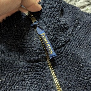 May include: Close-up of a navy blue fleece jacket with a gold zipper. The zipper pull features a small blue tag with the word "PENDLETON" in gold lettering. The jacket has a textured, fuzzy appearance.