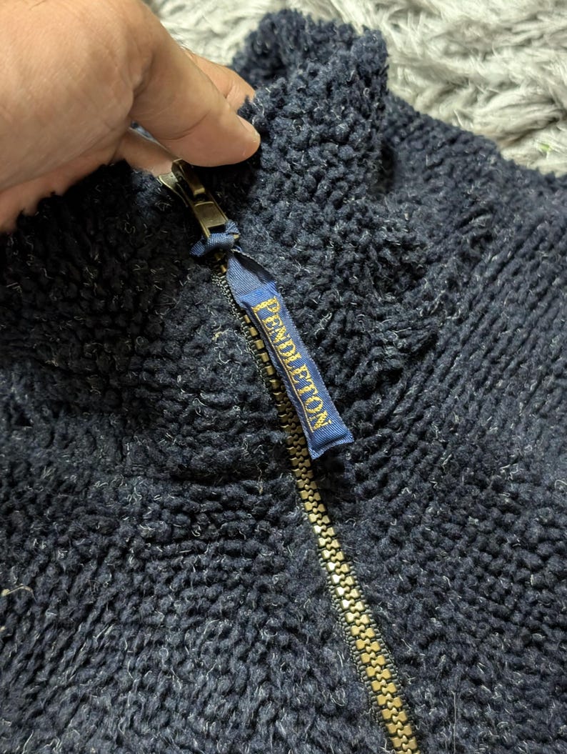 May include: Close-up of a navy blue fleece jacket with a gold zipper. The zipper pull features a small blue tag with the word "PENDLETON" in gold lettering. The jacket has a textured, fuzzy appearance.