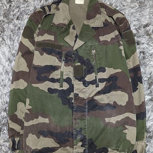 May include: A long-sleeve camouflage jacket featuring a woodland pattern in shades of green, brown, and black. The jacket includes a collared neckline, two vertical zippered chest pockets, shoulder epaulets, and circular patches on both sleeves.