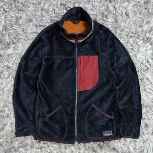 May include: A black fuzzy fleece jacket with a full-length front zipper, a stand-up collar, and a contrasting dark red chest pocket. The interior features an orange mesh lining, and a small rectangular label with text is on the lower front.