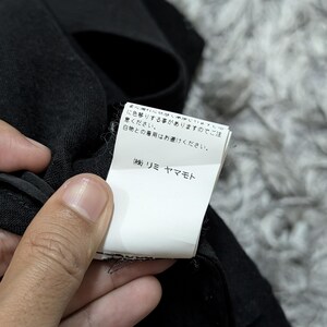 May include: A black garment featuring a white rectangular care label with Japanese text. The label includes the text: まに濡れた状態や摩擦により色移りする事がありますのでご注意ください。白物との着用はお避けください。 (株) リミ ヤマモト. The fabric has a textured, woven appearance.