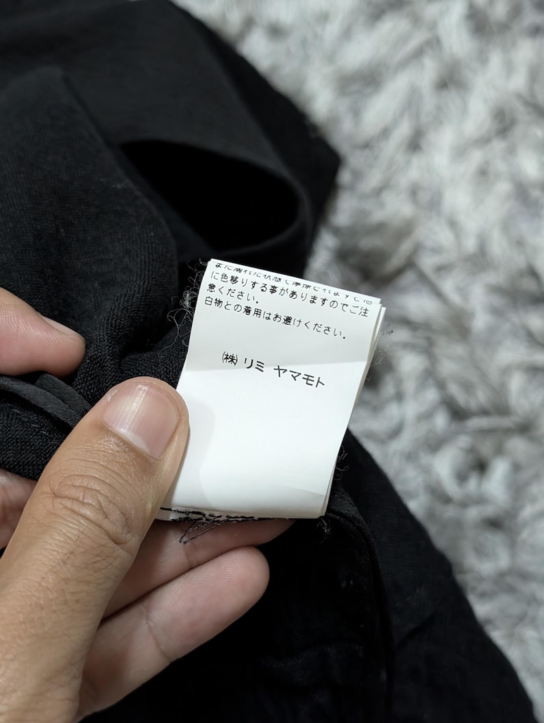 May include: A black garment featuring a white rectangular care label with Japanese text. The label includes the text: まに濡れた状態や摩擦により色移りする事がありますのでご注意ください。白物との着用はお避けください。 (株) リミ ヤマモト. The fabric has a textured, woven appearance.