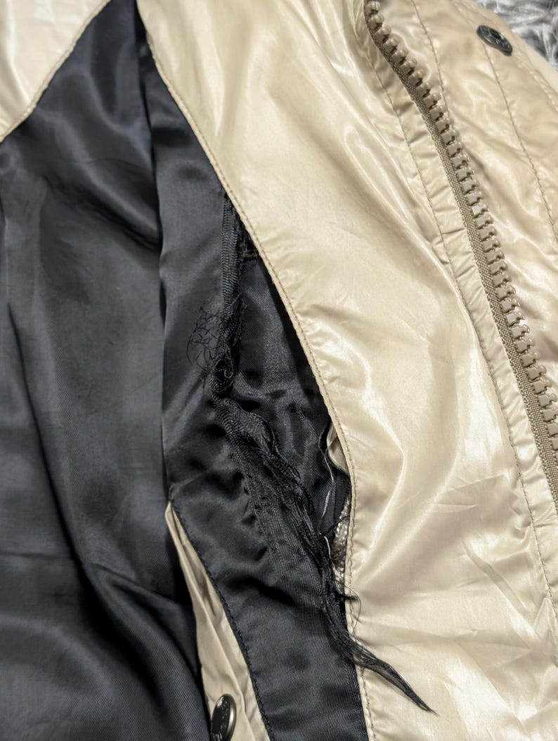Peut inclure: Gros plan sur une veste beige avec une doublure noire. La veste est dot&eacute;e d'une fermeture &eacute;clair et d'un bouton. La doublure est d&eacute;chir&eacute;e, r&eacute;v&eacute;lant la construction int&eacute;rieure.