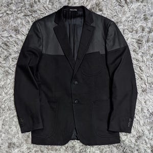 May include: A black blazer featuring a contrasting yoke panel across the chest and shoulders. This garment includes a notched lapel, two front buttons, a single chest pocket, two lower patch pockets, and four buttons on each sleeve cuff.