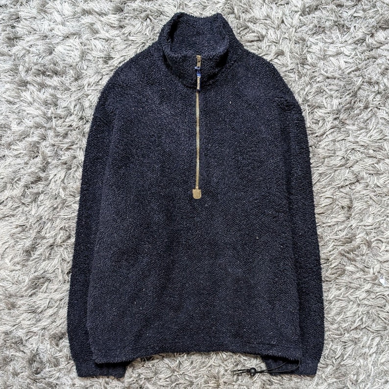 May include: A navy blue fleece pullover with a high collar and a gold-colored zipper. The long-sleeved top has a textured appearance and a drawstring at the bottom hem. The garment is displayed on a gray, textured surface.