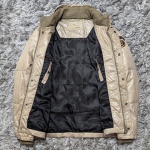 Peut inclure: Veste matelass&eacute;e beige avec une doublure noire. La veste a un col montant, plusieurs poches et une fermeture &eacute;clair sur le devant. Il y a un patch sur la manche sup&eacute;rieure droite et une &eacute;tiquette sur la poitrine. La veste est ouverte, r&eacute;v&eacute;lant l'int&eacute;rieur noir.