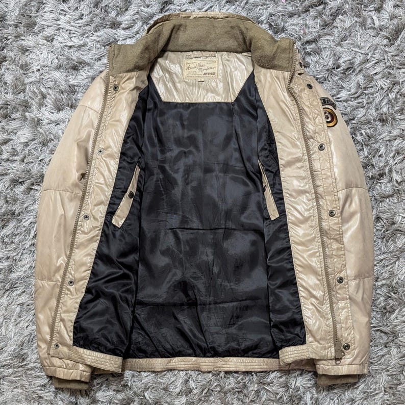 Peut inclure: Veste matelass&eacute;e beige avec une doublure noire. La veste a un col montant, plusieurs poches et une fermeture &eacute;clair sur le devant. Il y a un patch sur la manche sup&eacute;rieure droite et une &eacute;tiquette sur la poitrine. La veste est ouverte, r&eacute;v&eacute;lant l'int&eacute;rieur noir.