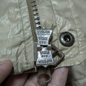 Peut inclure: Gros plan sur une fermeture &eacute;clair de veste beige avec le nom de marque "VISLON" et "YKK" visibles sur le curseur. La veste a une texture matelass&eacute;e et un bouton-pression en m&eacute;tal.