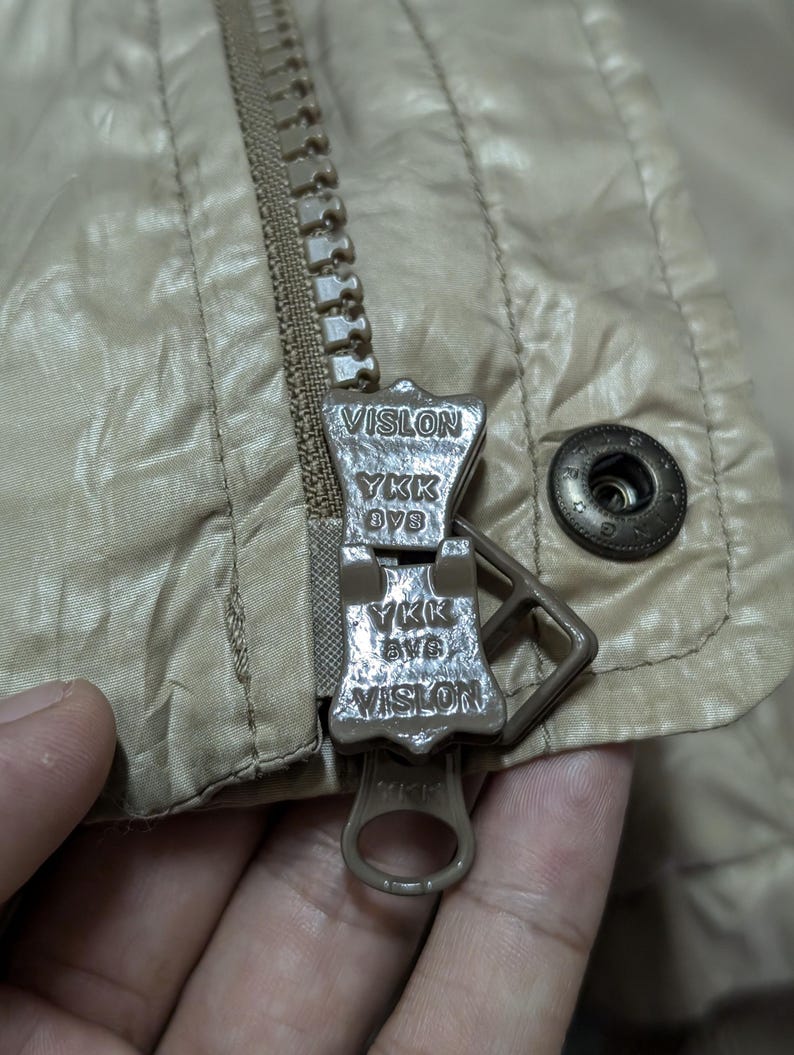 Peut inclure: Gros plan sur une fermeture &eacute;clair de veste beige avec le nom de marque "VISLON" et "YKK" visibles sur le curseur. La veste a une texture matelass&eacute;e et un bouton-pression en m&eacute;tal.