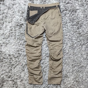 May include: Tan trousers featuring a folded-over waistband with a black and white polka dot pattern, side button closures, and a matching polka dot trim on the back pocket. The trousers have a gathered, textured appearance along the legs.