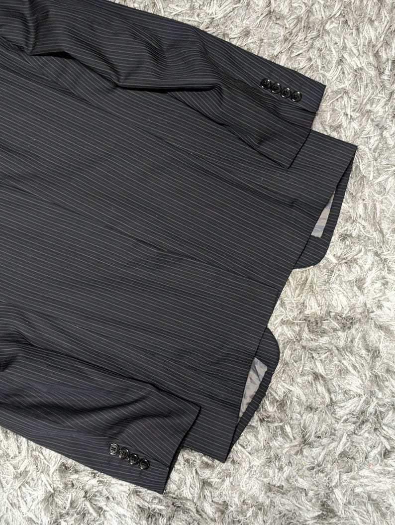 May include: A dark gray pinstripe blazer with four buttons on each sleeve. The blazer is made of a textured fabric and has a classic design. The interior lining is visible in the image.