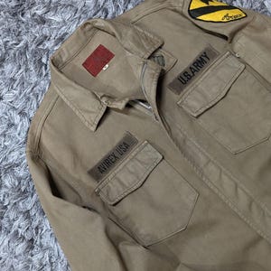 May include: A tan long-sleeve jacket with a zip fastening, a collar, and two chest pockets. The jacket features a red interior label, a yellow and black shoulder patch, and embroidered patches that read U.S. ARMY and AVIREX USA on the chest.