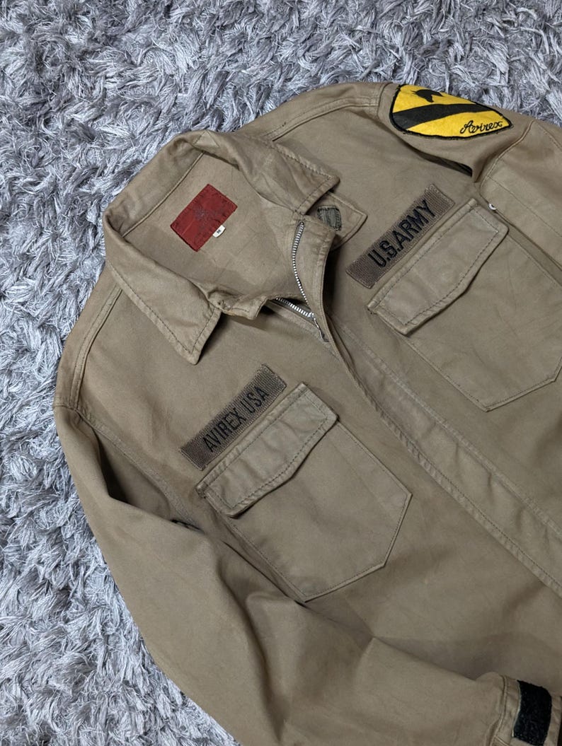 May include: A tan long-sleeve jacket with a zip fastening, a collar, and two chest pockets. The jacket features a red interior label, a yellow and black shoulder patch, and embroidered patches that read U.S. ARMY and AVIREX USA on the chest.