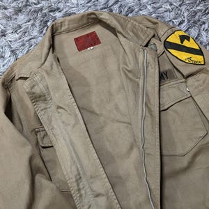 May include: A tan jacket featuring a front zip fastening, a collar, and two chest pockets. A red label is sewn inside the collar. A yellow and black patch with a horse silhouette and the text Avirex is on the shoulder, with an Army patch below.