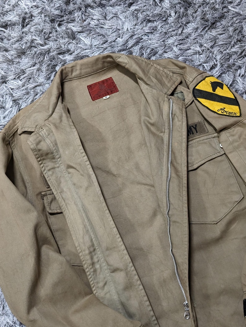 May include: A tan jacket featuring a front zip fastening, a collar, and two chest pockets. A red label is sewn inside the collar. A yellow and black patch with a horse silhouette and the text Avirex is on the shoulder, with an Army patch below.