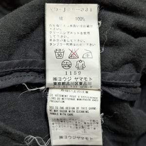 May include: A white rectangular garment care label stitched onto a dark gray fabric. The label features black text including 100% cotton, care instructions with symbols, and the company name Yohji Yamamoto, along with address and warning text.
