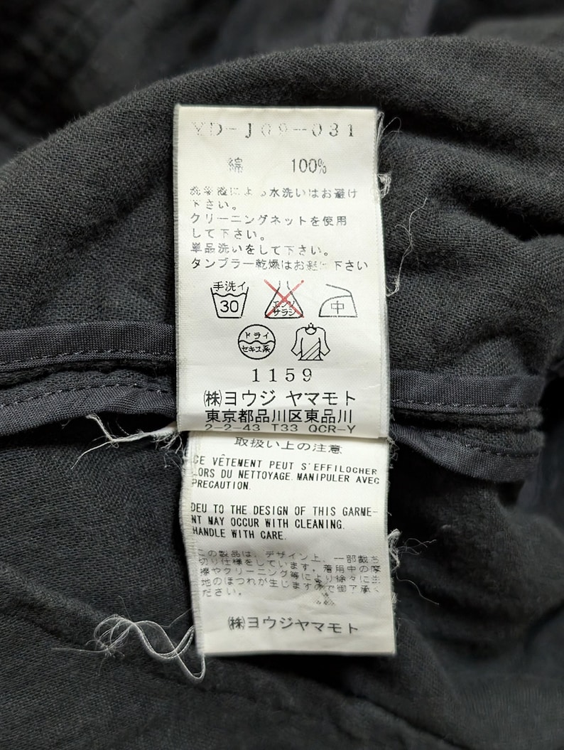 May include: A white rectangular garment care label stitched onto a dark gray fabric. The label features black text including 100% cotton, care instructions with symbols, and the company name Yohji Yamamoto, along with address and warning text.