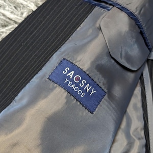 May include: Close-up of the interior of a pinstripe suit jacket. The lining is a light gray color, and a blue label with the text "SACSNY Y'SACCS" is sewn inside. The jacket's exterior is black with thin white stripes.