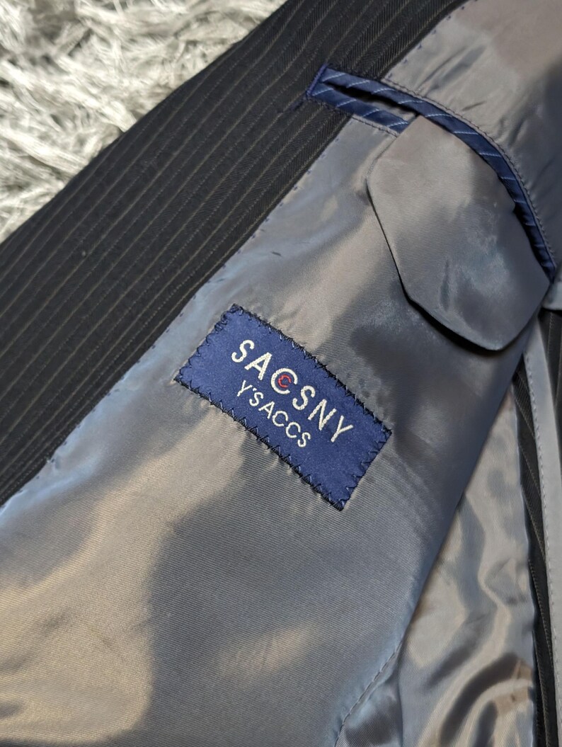 May include: Close-up of the interior of a pinstripe suit jacket. The lining is a light gray color, and a blue label with the text "SACSNY Y'SACCS" is sewn inside. The jacket's exterior is black with thin white stripes.