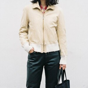 May include: A beige jacket with a full-length front zip, a pointed collar, and ribbed white cuffs at the sleeves and hem. The jacket is worn over a patterned shirt, paired with dark trousers and a black tote bag held at the side.
