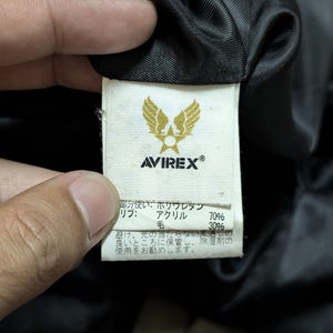 Peut inclure: Gros plan sur une &eacute;tiquette blanche avec le logo "AVIREX" et un embl&egrave;me dor&eacute; d'ailes. L'&eacute;tiquette contient du texte japonais d&eacute;taillant la composition du tissu : polyur&eacute;thane, acrylique (70%) et laine (30%).