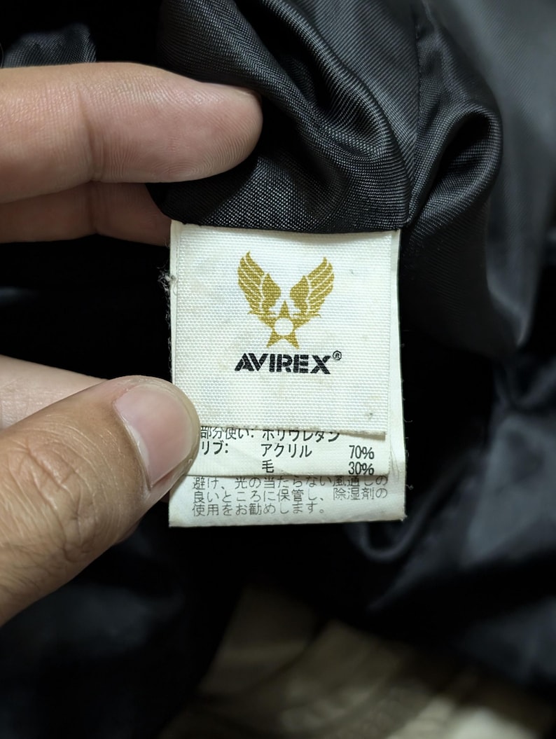 Peut inclure: Gros plan sur une &eacute;tiquette blanche avec le logo "AVIREX" et un embl&egrave;me dor&eacute; d'ailes. L'&eacute;tiquette contient du texte japonais d&eacute;taillant la composition du tissu : polyur&eacute;thane, acrylique (70%) et laine (30%).