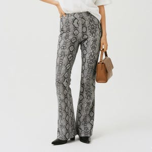 Puede incluir: Pantalones de talle alto con estampado de piel de serpiente en gris y negro y pernera acampanada. Los pantalones se combinan con un bolso estructurado de color marrón liso con asa superior y cierre de solapa, junto a una camiseta blanca.