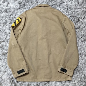 May include: A tan long-sleeve jacket featuring a fold-down collar and a yellow and black patch on the right shoulder. The cuffs are adjustable with black hook-and-loop fasteners. The jacket is constructed from a solid-coloured, durable fabric.