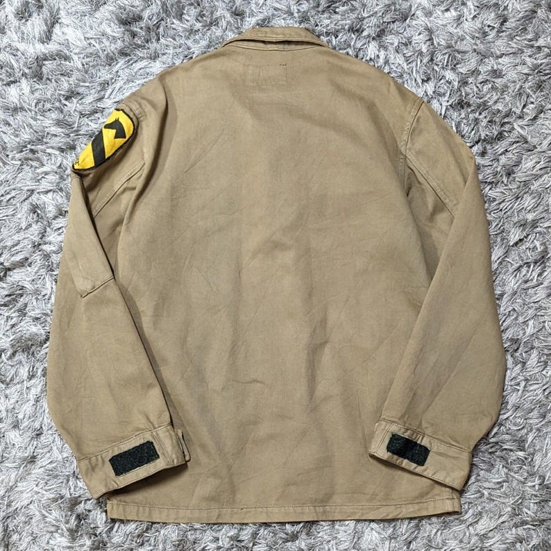 May include: A tan long-sleeve jacket featuring a fold-down collar and a yellow and black patch on the right shoulder. The cuffs are adjustable with black hook-and-loop fasteners. The jacket is constructed from a solid-coloured, durable fabric.