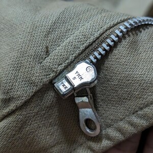 May include: A tan-coloured fabric garment featuring a silver-toned metal zip closure. The zip pull and slider are clearly marked with the YKK brand logo and the number 5, indicating the size of the zip track and the manufacturer's mark.