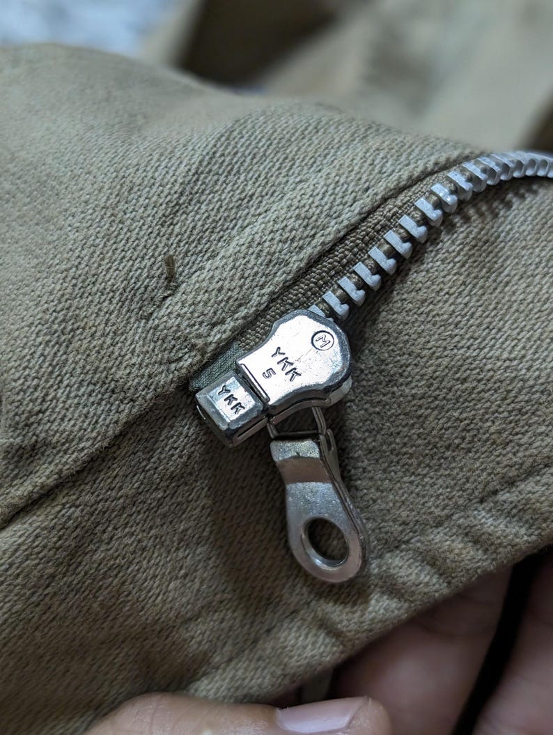 May include: A tan-coloured fabric garment featuring a silver-toned metal zip closure. The zip pull and slider are clearly marked with the YKK brand logo and the number 5, indicating the size of the zip track and the manufacturer's mark.
