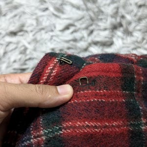 May include: Close-up of a section of a red, green, and white plaid fabric. The fabric has a textured appearance and is held by a hand. Two metal clasps are visible on the fabric. The background is a blurred, light-colored surface.