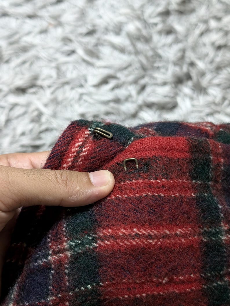 May include: Close-up of a section of a red, green, and white plaid fabric. The fabric has a textured appearance and is held by a hand. Two metal clasps are visible on the fabric. The background is a blurred, light-colored surface.