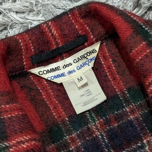May include: Close-up of a red, green, and navy plaid wool garment. The label reads "COMME des GAR&Ccedil;ONS" and "M". The garment is made of 100% wool and made in Italy.