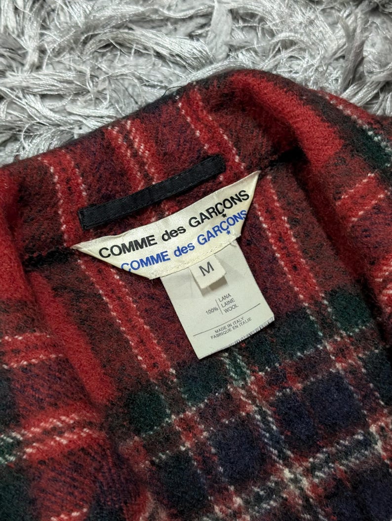 May include: Close-up of a red, green, and navy plaid wool garment. The label reads "COMME des GAR&Ccedil;ONS" and "M". The garment is made of 100% wool and made in Italy.