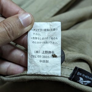 May include: A white rectangular garment care label attached to a tan fabric item. The label features Japanese text, the company name Ueno Shokai, a phone number 03-3831-1546, and the text 中国製. A dark brown button is visible behind the label.