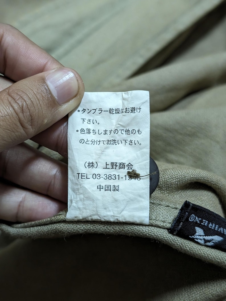 May include: A white rectangular garment care label attached to a tan fabric item. The label features Japanese text, the company name Ueno Shokai, a phone number 03-3831-1546, and the text 中国製. A dark brown button is visible behind the label.