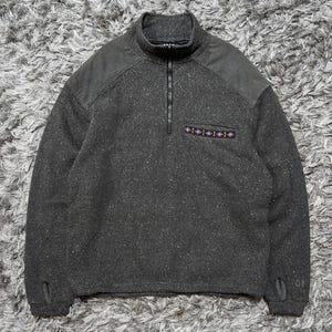 Puede incluir: Un suéter de forro polar gris carbón moteado con cuello de cremallera corta, paneles de hombro en gris oscuro liso y un bolsillo en el pecho con un adorno geométrico. Las mangas largas incluyen orificios para los pulgares en los puños.